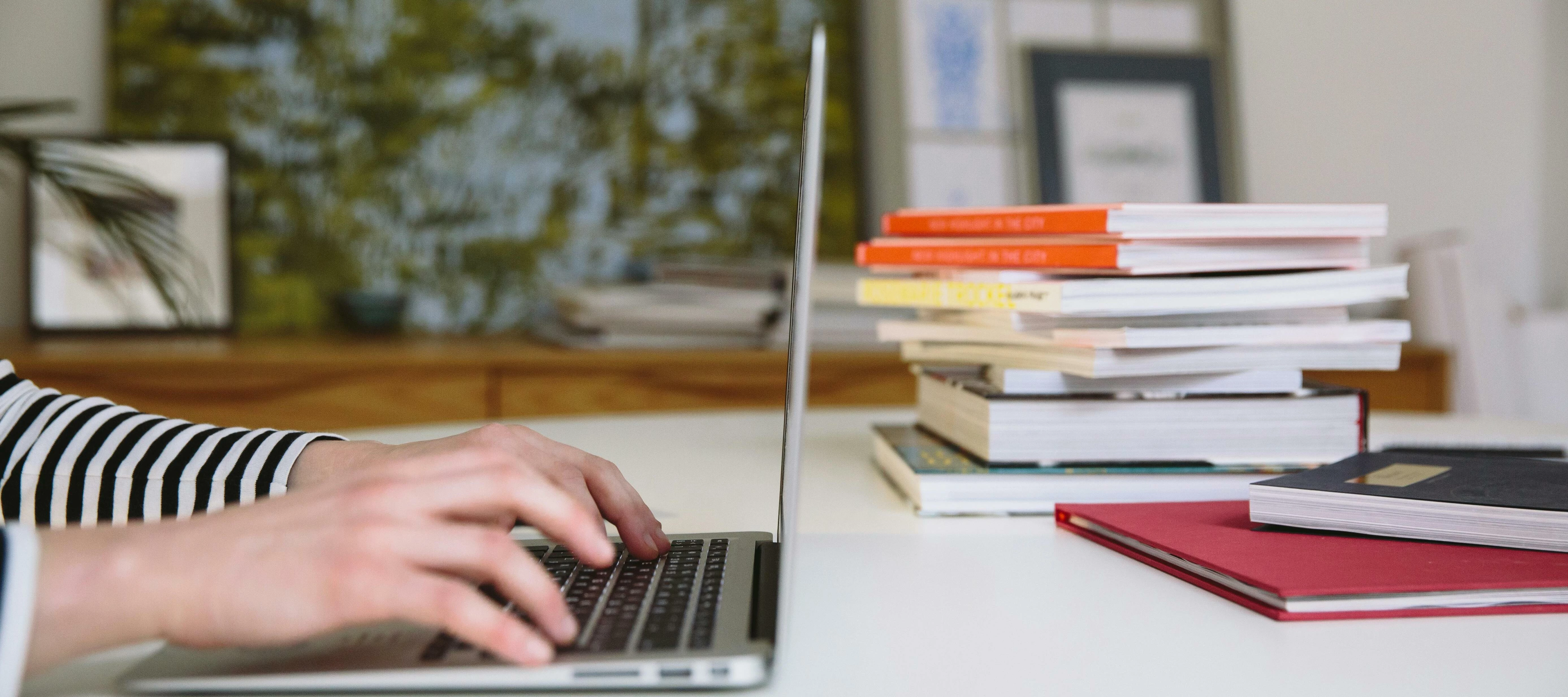 person typing on a laptop with books stacked up in the background