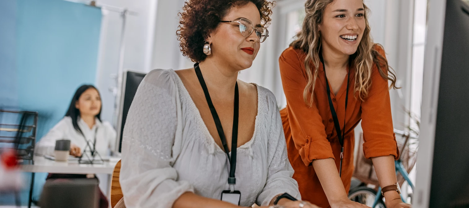 Two women sit at a desk looking towards a computer. One is hovering over the other.
