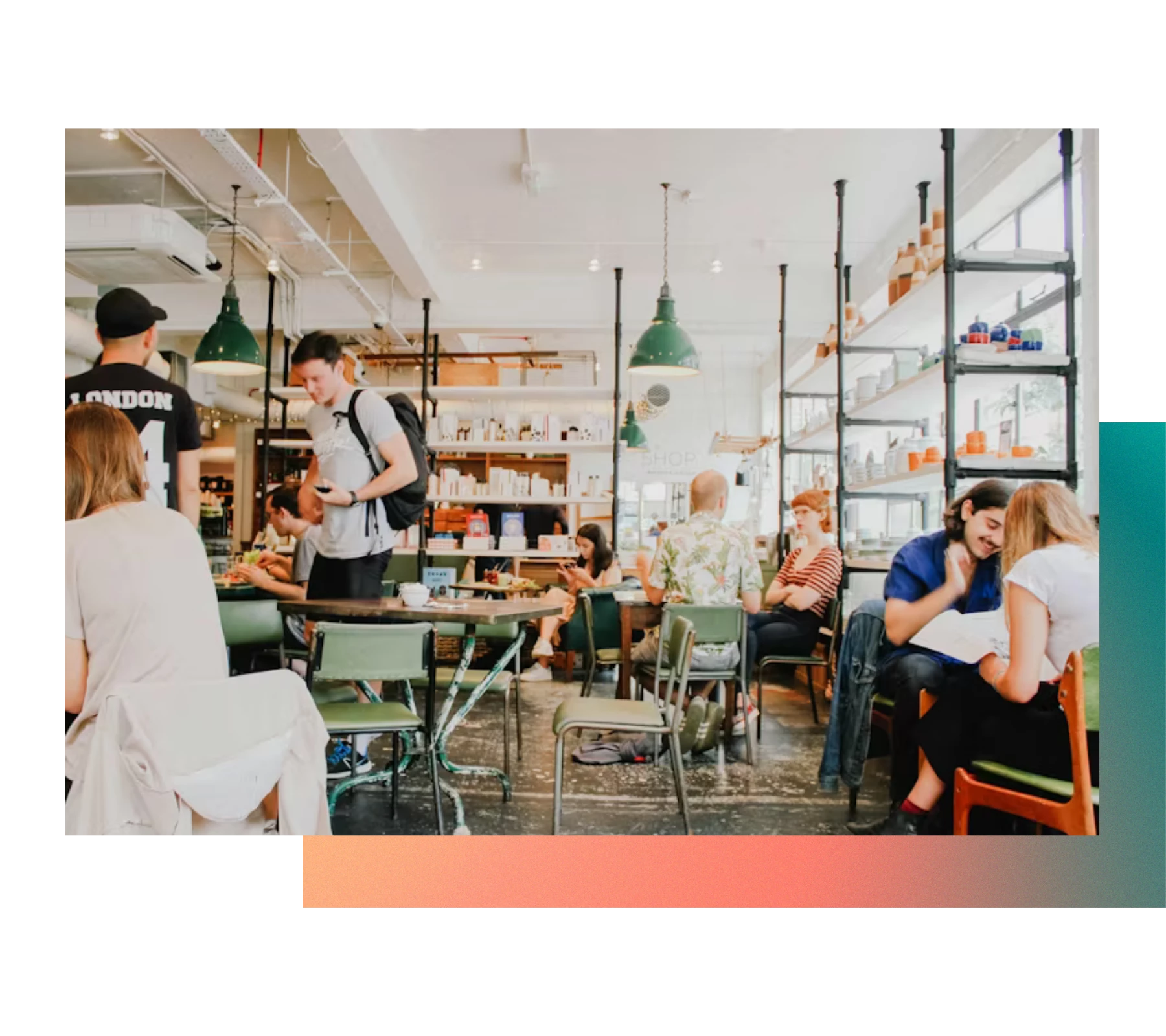 A bustling café with several customers seated at tables, engaged in conversations. One person is standing with a backpack, looking at a mobile device. The space is bright, with shelves lined with various items and large, green hanging lights.