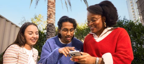 Three people gather together to look at something on a cell phone together. 